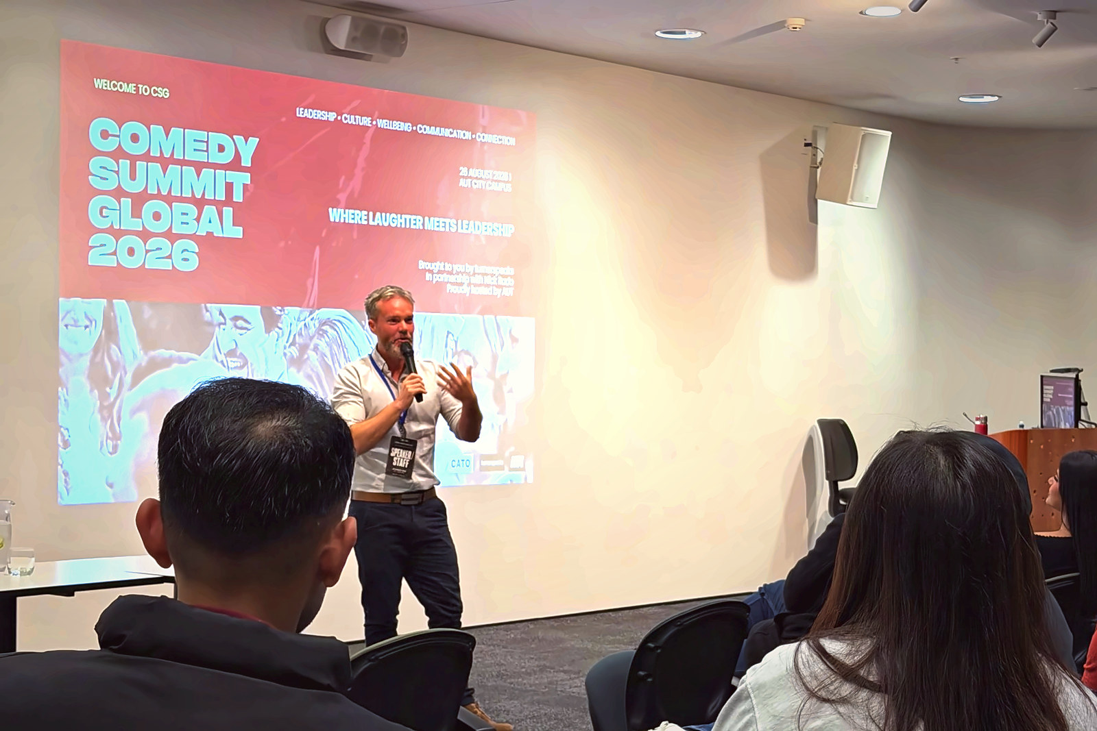 A speaker presenting on stage beside the Comedy Summit Global 2026 title slide at AUT